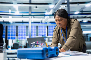 African american woman consultant in solar energy reviews data on laptop, inspecting production line and manufacturing system. Industrial employee oversees plant performance for innovation.
