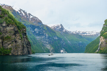 Boats float within foliage on rocky mountain cliffs, snowcapped mountains, and waterfalls of Norway's most gorgeous UNESCO designated fjord, Geirangerfjord