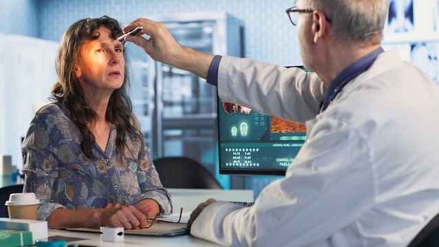 Male old healthcare provider conducts a concussion test in a medical clinic, evaluating the woman patient focus, coordination and pupil reaction after a suspected head trauma. Camera B.