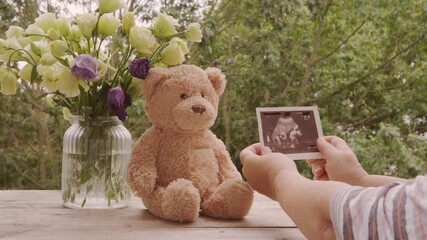 Ultrasound announcement card, sonogram image in hand, flowers in glass vase, Teddy bear on wooden table, pregnancy reveal, motherhood, baby coming soon, prenatal care, family joy, waiting for baby