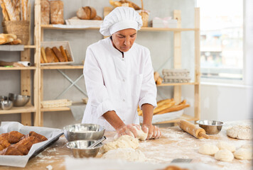 Positive senior female baker in white uniform kneading dough and preparing dough for baking at light kitchen. Bakery work