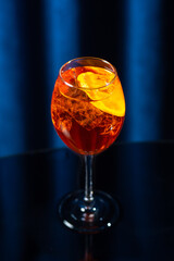 A glass of orange cocktail with ice cubes and orange slices on a dark glossy surface with a blue curtain background