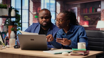 Mature couple dealing with online banking together at home in their loft, setting up payments and confirming money transfer. Pleased man and woman enjoy online shopping on laptop. Camera B.