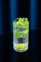 A close-up of a mojito cocktail in a tall crystal glass with mint leaves, lime slices, and ice, set against a dark blue background