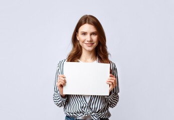 Naklejka premium Portrait of beautiful woman holding white blank advertising board, smiling millennial lady demonstrating empty banner with copy space for your text or design, standing on gray background in studio