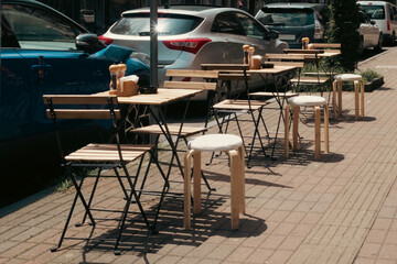 Wooden tables and chairs of a street cafe line the sunny sidewalk in Batumi, Georgia. Urban summer atmosphere with parked cars in the background.
