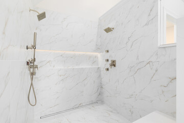 A large walk-in shower with large format white marble tiled walls, gold faucets, and a shelf with...
