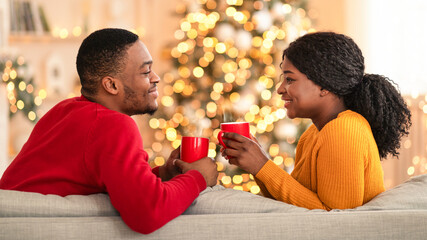New year mood at home and romantic celebration together. Smiling millennial african american couple with cups with hot drink in interior of living room with Christmas tree and glowing garlands