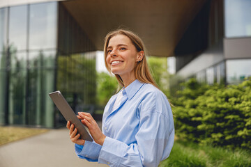 Fototapeta premium A smiling young woman confidently using a tablet outdoors next to a contemporary building in the city