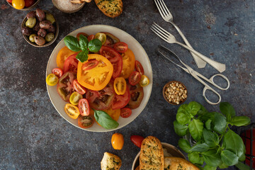 Overhead view of mixed tomatoes, basil stems, olives, salt bowl and pepper mill arranged diagonally on dark background