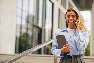 A cheerful young woman dressed in professional business attire is talking on the phone while holding a tablet outside