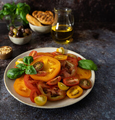 Front view of colorful sliced tomatoes with basil, olive oil bottle, bread and olives on dark blue brown background
