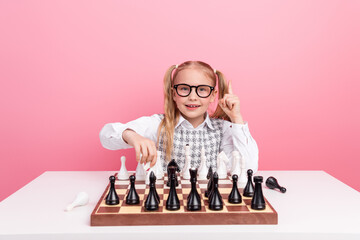 Smart young girl in glasses playing chess with focus and enthusiasm on a pink background, learning strategy