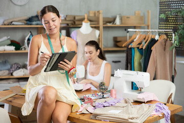 Dressmaker comes up with a design for a clothing collection and draws it on a tablet, she works against the background of a seamstress in the workshop. Team of a fashion designer and a dressmaker work