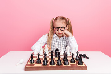 Thoughtful young girl playing chess at a table with a pink background, showcasing focus and strategic thinking