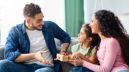 Portrait of cute little daughter and wife making surprise for arab man, giving present box at home, greeting happy guy with Father's day or birthday, joyful family bonding on couch in living room
