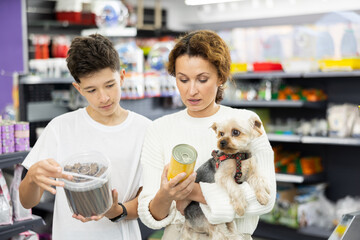 Interested woman accompanied by teenage son meticulously selecting quality dog food for beloved Yorkshire terrier in pet store, attentively examining packaging and considering choice