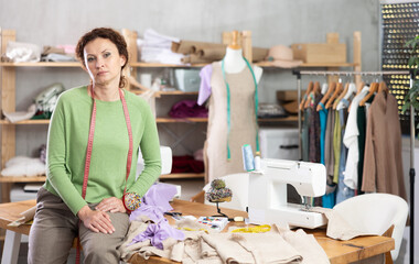 Professional female dressmaker with measuring tape around neck sitting relaxed on edge of work...