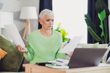 Serious woman analyzing documents at home while working on her computer, wearing a casual green jumper, in a stylish living space.