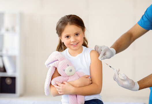 Happy little girl with toy bunny getting coronavirus vaccine injection at hospital. Black doctor immunizing child against covid-19 virus at clinic. Healthcare and medical concept - Powered by Adobe