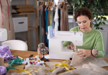 Focused female seamstress operating sewing machine in workshop filled with fabrics, carefully working on bespoke linen garment surrounded by tools