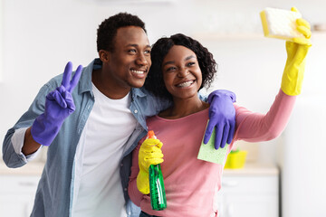 Cheerful african american lovers having fun while cleaning kitchen, holding spray bottles and wearing rubber gloves, funny black man and woman taking selfie, using sponge as smartphone