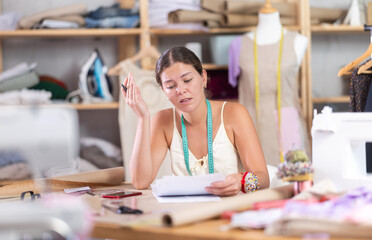 Upset young tailoress struggling with paperwork sitting at table in fashion studio