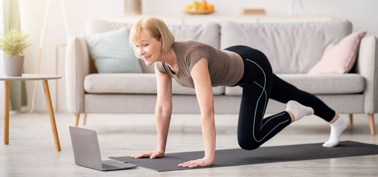 Online sports during home isolation. Fit mature lady doing exercises in front of laptop indoors, copy space. Strong senior woman following video tutorial, working out on yoga mat, training her muscles