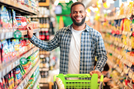 Happy African American Guy Posing In Supermarket. Cheerful Customer Choosing Food And Buying Groceries Standing Near Shelf In Local Store Indoors, Smiling To Camera. Selective Focus - Powered by Adobe