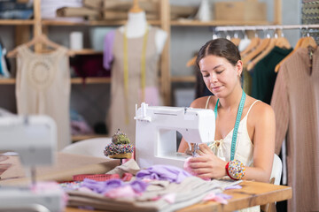 Positive young female dressmaker working with sewing machine in tailor's workshop