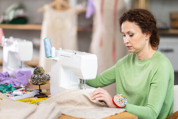 Dedicated female tailor fulfilling custom garment from linen fabric using modern sewing machine in fashion studio filled with materials and tools