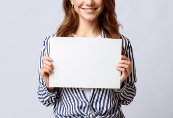 Naklejka premium Unrecognizable young lady holding blank sheet of paper in hands, smiling woman demonstrating empty placard with copy space for your text or design, posing over light background, closeup view