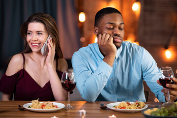 Bad Date. Black guy sitting bored while his girlfriend talking on cellphone in restaurant, ignoring him during romantic Valentine's day dinner. Multiracial couple having problems in relationship