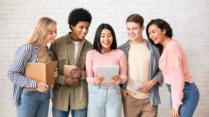 Interesting site, new app for study, test results online and teamwork. Smiling young multiracial people with books and gadgets look at tablet of asian lady in classroom, studio shot, empty space