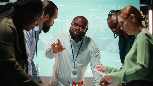 Night time meeting unfolds as a multiethnic team gathers in a strategy room, analyzing visual data, statistics and risk assessment files to drive project development for efficiency. Camera B.