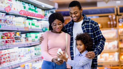 Cute black little girl with her parents holding bottle of milk, shopping together, buying dairy products at supermarket. Millennial African American family purchasing groceries at mall on weekend
