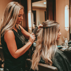 Fototapeta premium Hair stylist brushing long blonde hair of female client in salon with warm lighting