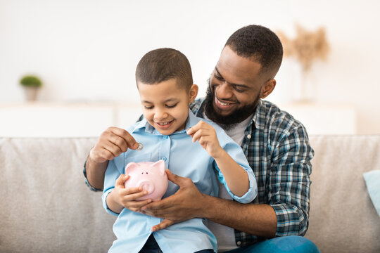 Financial Literacy. Cheerful African Father And Son Putting Money In Piggybank Sitting On Sofa At Home. Daddy Teaching His Child Budget Planning, Keeping Personal Savings Safety - Powered by Adobe