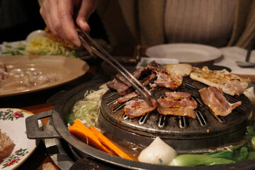 Grilled meats cooking on a Korean barbecue pan with a hand using tongs, surrounded by fresh vegetables and dipping sauces on a restaurant table.