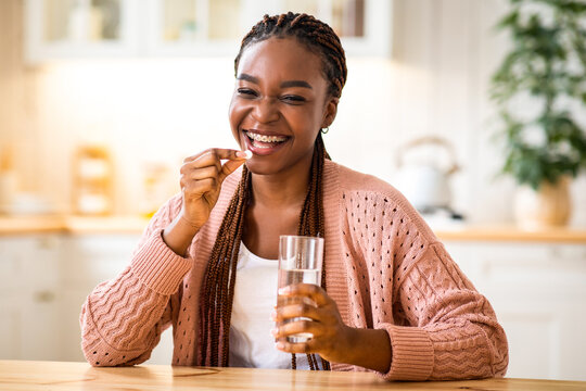 Beauty Supplements. Positive Young Black Female Taking Vitamin Pill And Drinking Water At Home, Smiling African Lady Using Multivitamin Tablet For Beautiful Skin, Hair And Nails, Looking At Camera