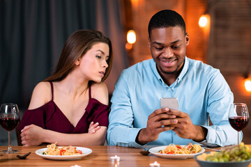 Jealous young woman peeking into boyfriend's smartphone, reading his messages during date dinner in restaurant, young black man having phubbing problem and ignoring his girlfriend, free space