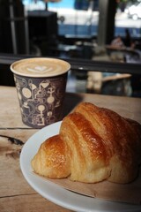 Fresh buttery croissant on a white plate next to a cup of latte with heart-shaped latte art, captured in a cozy café setting.