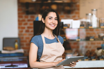 Opening small business. Happy arab woman in apron near bar counter holding digital tablet and looking at camera, waiting for clients in modern loft cafe