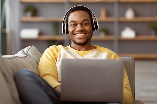 Joyful black guy reclining on couch at home, watching movie on laptop, using headset, copy space. Happy african american man playing video games or having video call while resting on weekend, closeup - Powered by Adobe