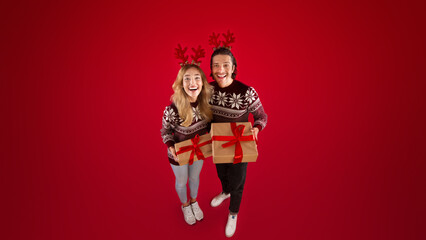 Happy couple wearing woolen sweaters and deer horns with Christmas gift over red background