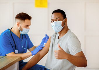 African American man in antiviral mask gesturing thumb up during coronavirus vaccination, approving of covid-19 immunization. Young doctor giving vaccine injection to male patient