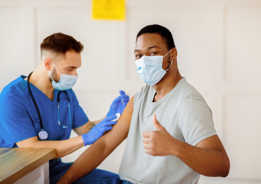 African American man in antiviral mask gesturing thumb up during coronavirus vaccination, approving of covid-19 immunization. Young doctor giving vaccine injection to male patient