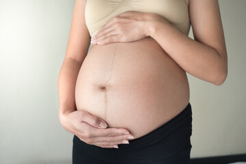 Pregnant asian woman gently holding belly and standing indoors. Soft smooth color tones at front view. Look warmth, care and motherhood. Concept of pregnancy, maternity, prenatal care, family planning