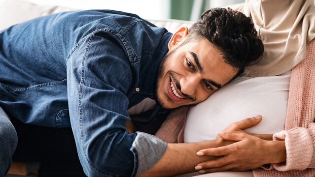 Happy Father. Joyful middle eastern man listening his pregnant wife's tummy, young arab guy tenderly hugging belly of expectant spouse, trying to feel baby kicks, cropped image, closeup - Powered by Adobe