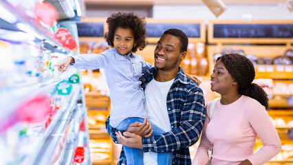 Happy black parents and their pretty daughter with shopping cart having fun buying groceries at hypermarket. African American family purchasing dairy products at modern mall